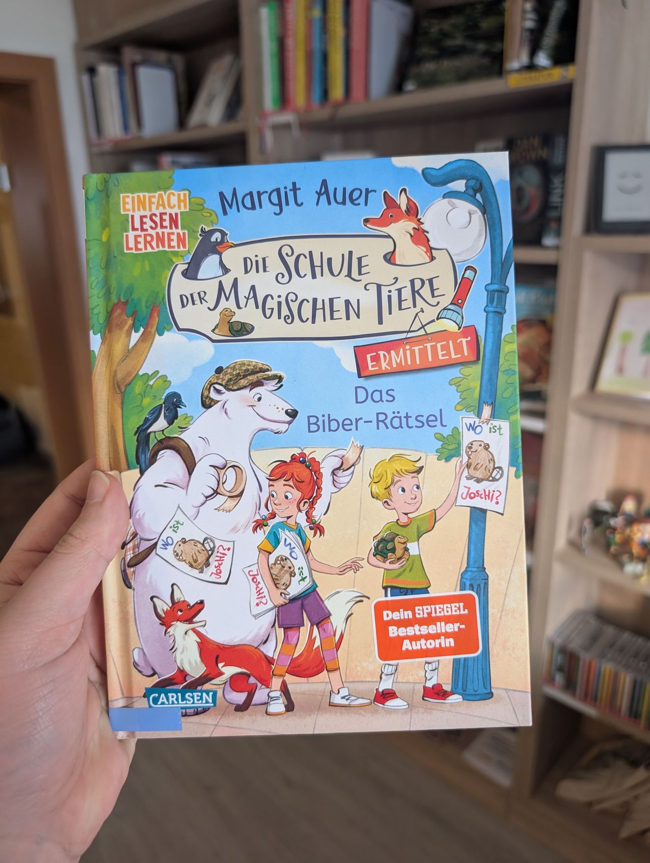 Ein Buch mit dem Titel 'Die Schule der Magischen Tiere' wird von einer Hand gehalten und zeigt einen Jungen, ein Mädchen und Tiere. Das Buchcover zeigt einen Fuchs, einen Bären und einen Pinguin.
