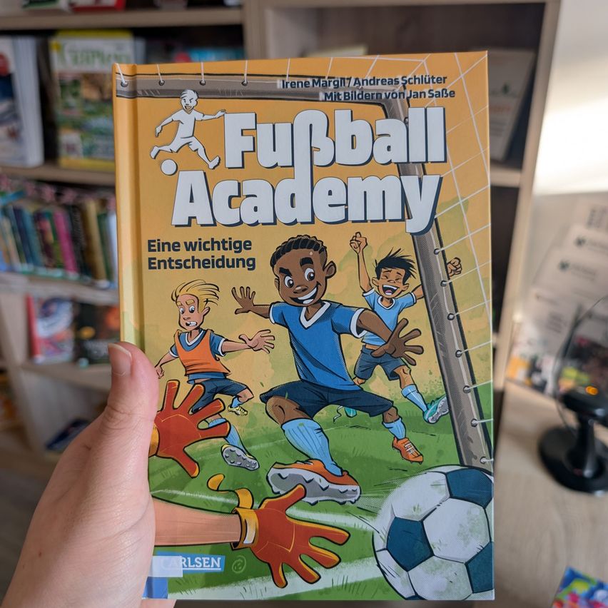 Eine Hand hält ein Buch mit dem Titel 'Fussball Academy' mit einem Fußball und Spielern auf dem Cover vor einem Bücherregal.