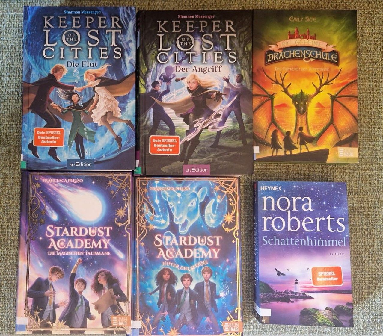 Sechs Bücher ausgestellt: zwei mit dem Titel 'Keeper of the Lost Cities', zwei mit dem Titel 'Stardust Academy' und zwei weitere. Die Bücher haben farbenfrohe Cover, die verschiedene Fantasieszenen darstellen.