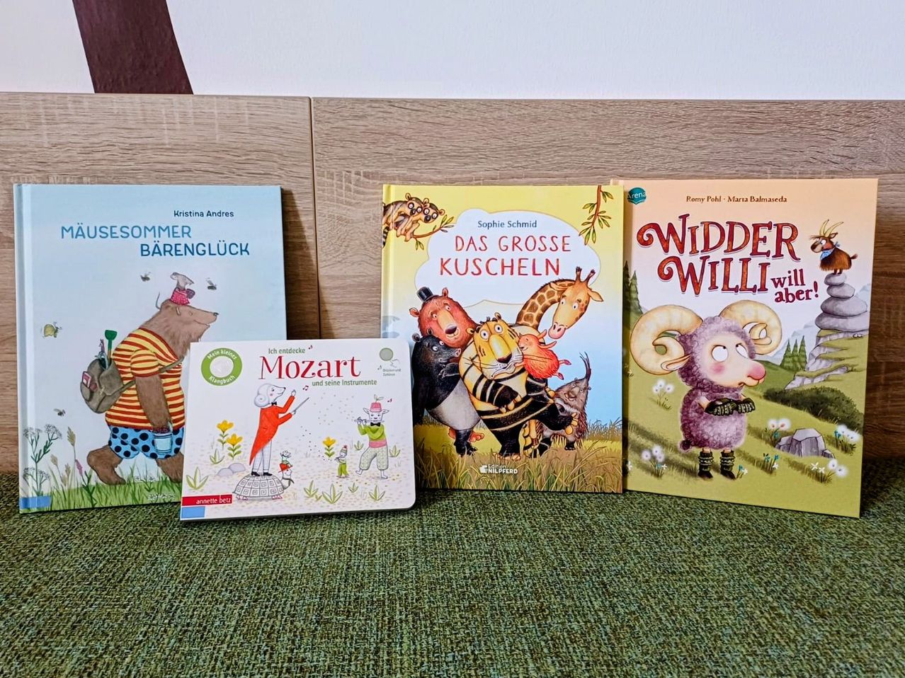 Vier Kinderbücher sind auf einem Holzregal ausgestellt. Die Bücher sind betitelt 'Sommer Barenglück', 'Das Grosse Kuscheln', 'Mozart' und 'Widde Willi'. Die Bücher sind bunt und haben zeichentrickähnliche Illustrationen. Die Bücher stehen auf einem grünen Teppich.