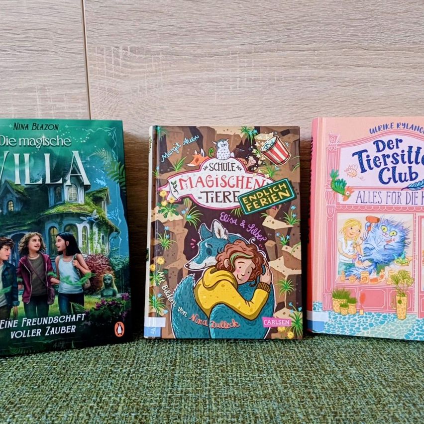 Drei Kinderbücher sind auf einer Holzoberfläche ausgestellt. Das linke Buch hat einen grünen Einband mit dem Titel 'Die magische Villa'. Das mittlere Buch hat einen braunen Einband mit dem Titel 'Die Schule der magischen Tiere'. Das rechte Buch hat einen rosa Einband mit dem Titel 'Der Tiersitter Club'.