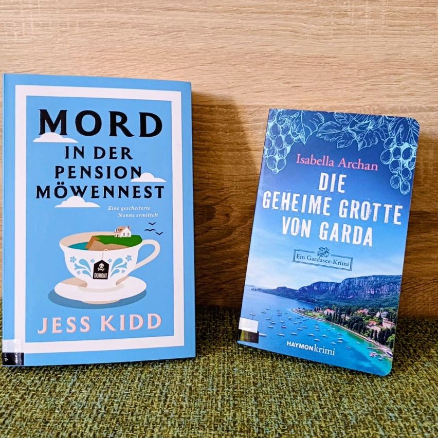 Zwei Bücher auf einer Holzoberfläche, eines mit dem Titel 'Mord in der Pension Möwennest' von Jess Kidd, das andere 'Die geheime Grotte von Garda' von Isabella Archan.