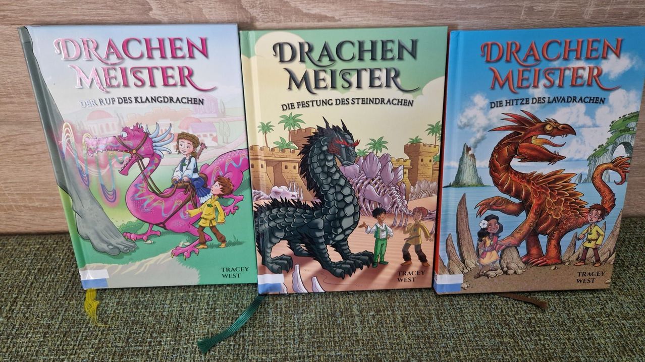 Three books titled Drachen Meister, Drachen Meister, and Drachen Meister are displayed on a wooden surface. The middle book features a dragon and a castle in the background.