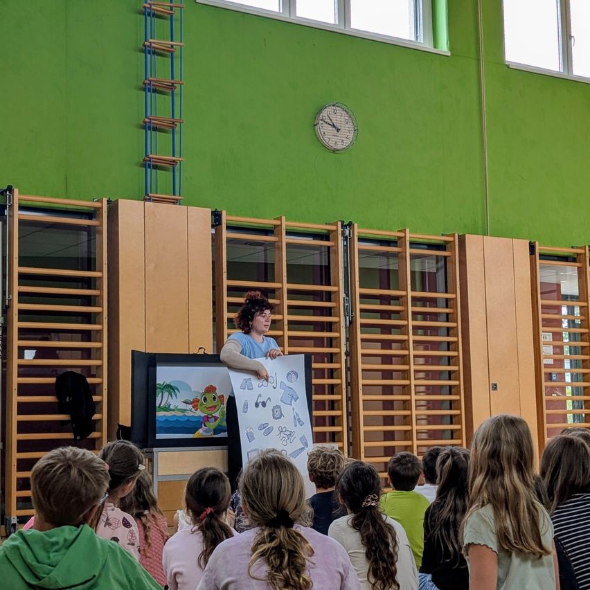 A teacher in a classroom holds up a board with a drawing while children sit in front of her. The room is green with large windows and a clock on the wall.