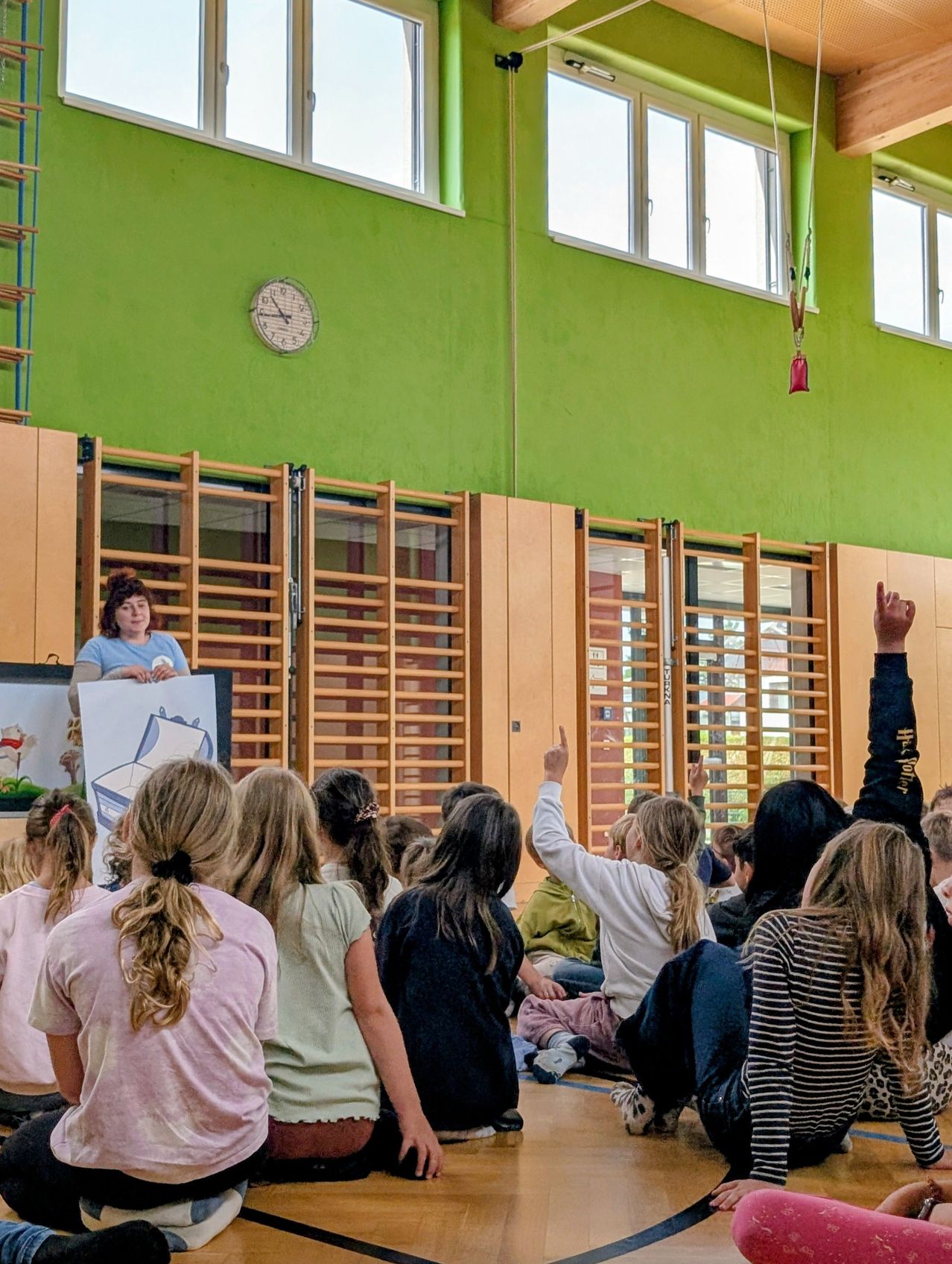 A classroom with green walls. A woman stands in front of the children. The children are sitting on the floor, some raising their hands. There is a clock and a red object hanging on the wall.