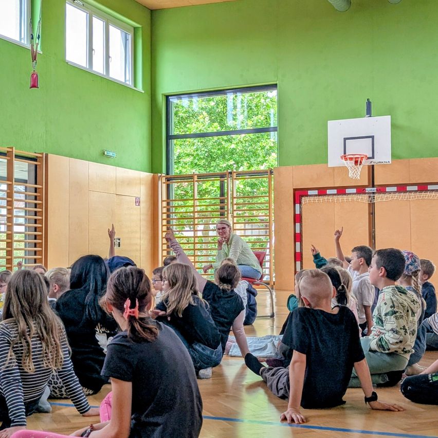 Inside a gymnasium, a woman addresses a seated group of children with raised hands. The room has green walls and a basketball hoop in the background.