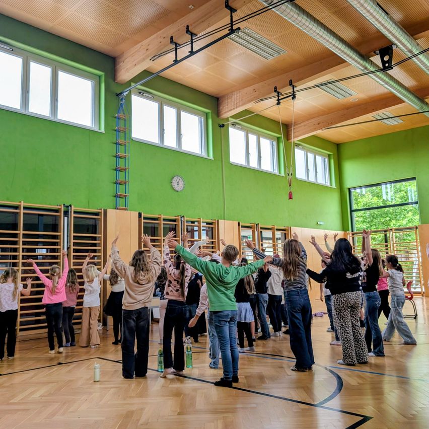 A group of people are performing an exercise routine inside a gymnasium with green walls and wooden floors. The room is well-lit with natural light from windows.