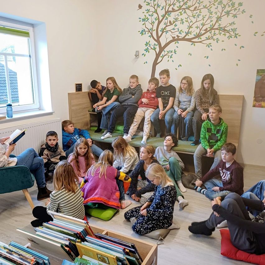 Children sit on the floor and on cushions, listening to a person read. A tree mural is on the wall.