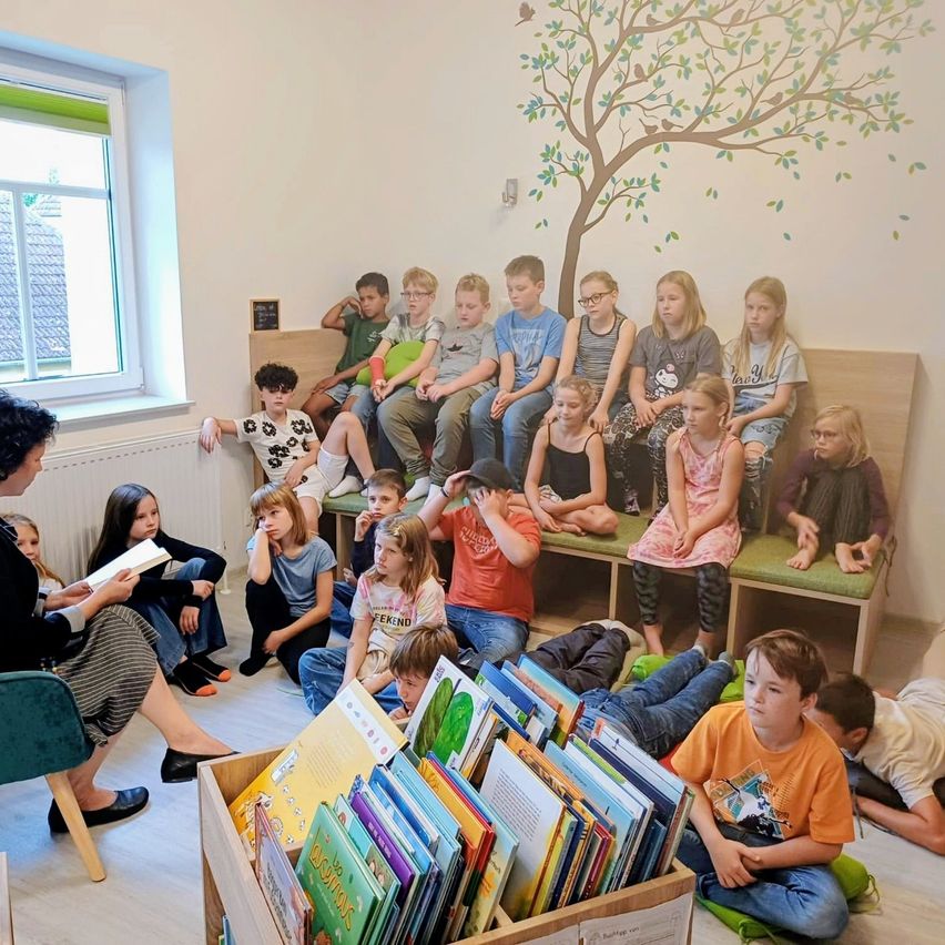 A group of children and a woman are sitting on benches and on the floor, reading books in a room with a window and a mural.