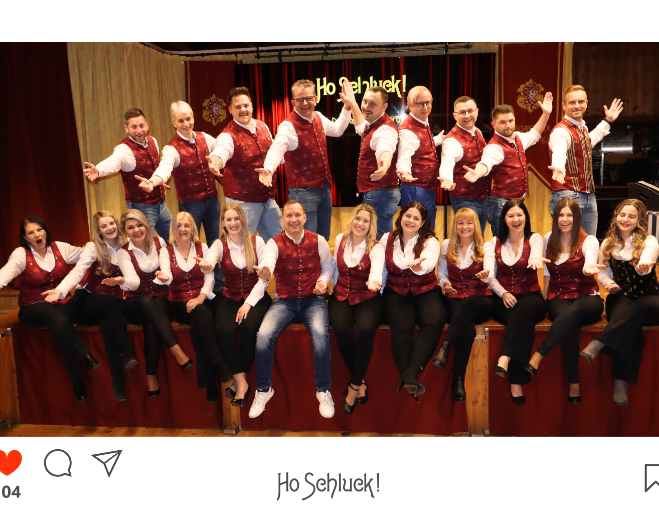 Eine Gruppe von Darstellern, die rote Westen tragen, posiert für ein Foto auf der Bühne, mit einem lächelnden Gesicht in der unteren linken Ecke.