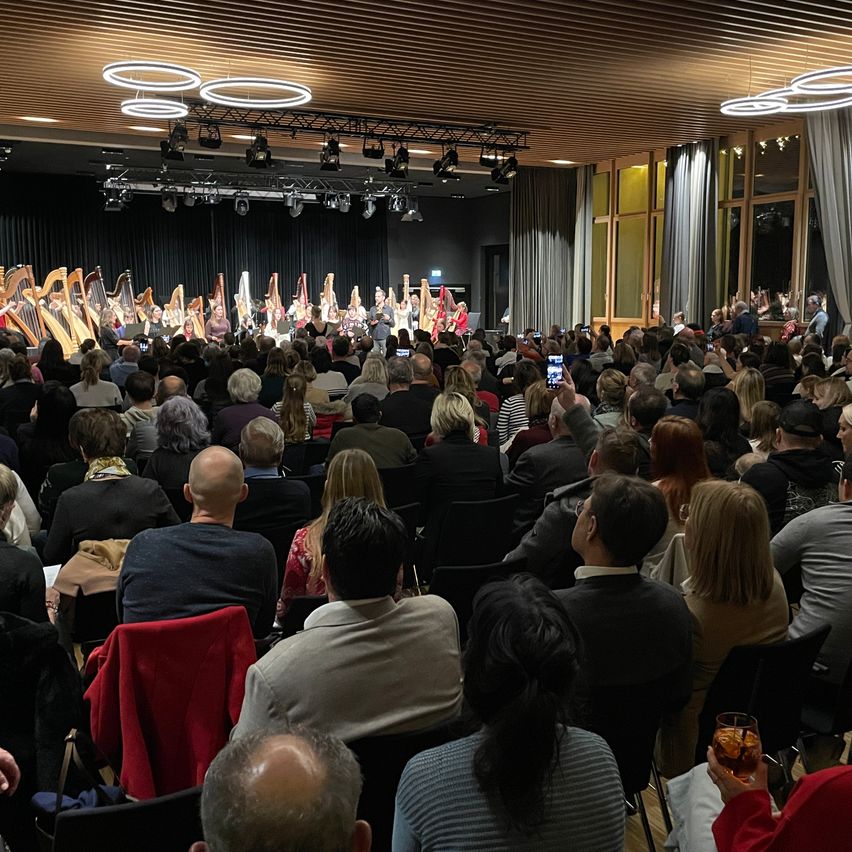 Ein großes Publikum sieht eine Harfenensemble-Aufführung auf der Bühne, einige halten das Moment mit ihren Handys fest.