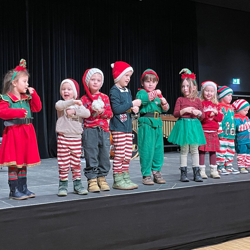 Kinder in Weihnachtskostümen, einschließlich Elfen und Weihnachtsmützen, treten auf einer Bühne mit schwarzem Hintergrund auf. Einige halten Musikinstrumente.