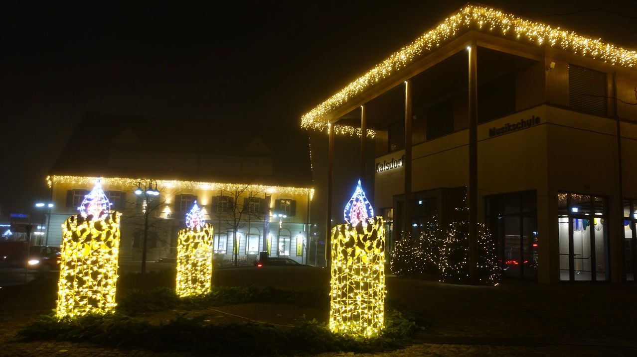 Zwei beleuchtete Kerzenskulpturen stehen bei Nacht vor einem Gebäude. Das Gebäude hat ein Schild mit der Aufschrift Kalsdorf. Das Gebiet ist mit Weihnachtslichtern geschmückt.