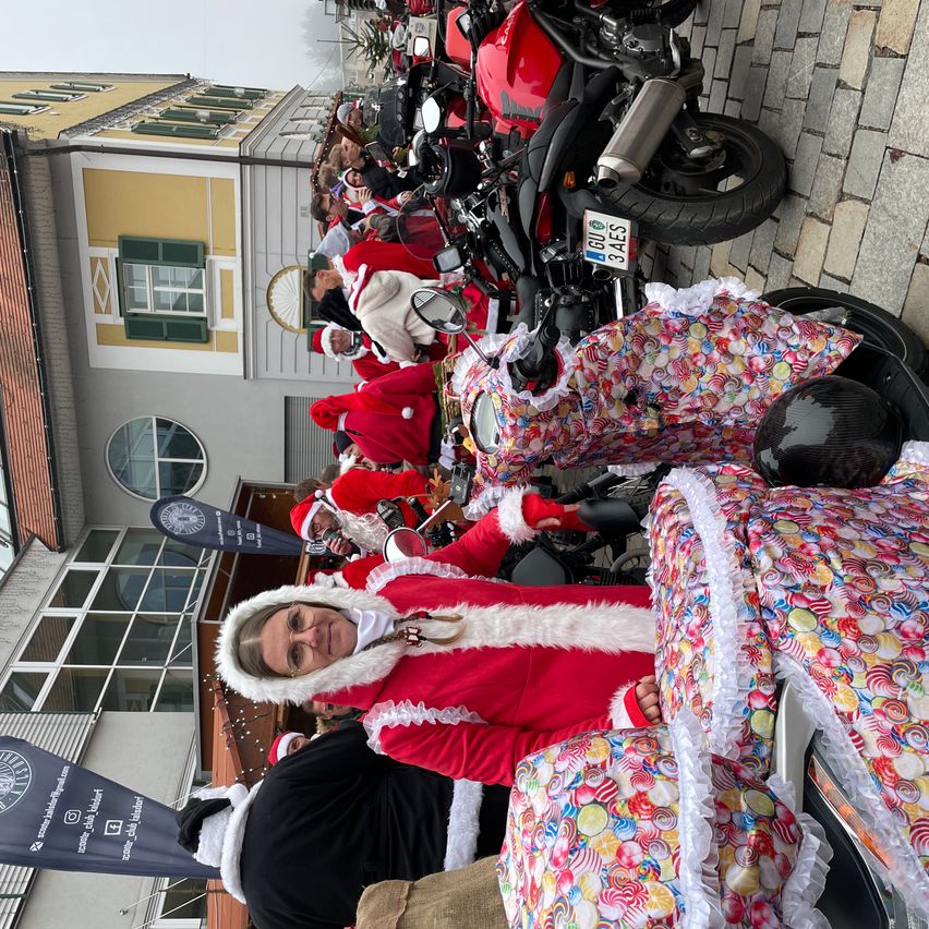 Eine Gruppe von Menschen in Weihnachtsmannkostümen versammelt sich um Motorräder vor einem Gebäude mit mehreren Fenstern und einer Fahne.