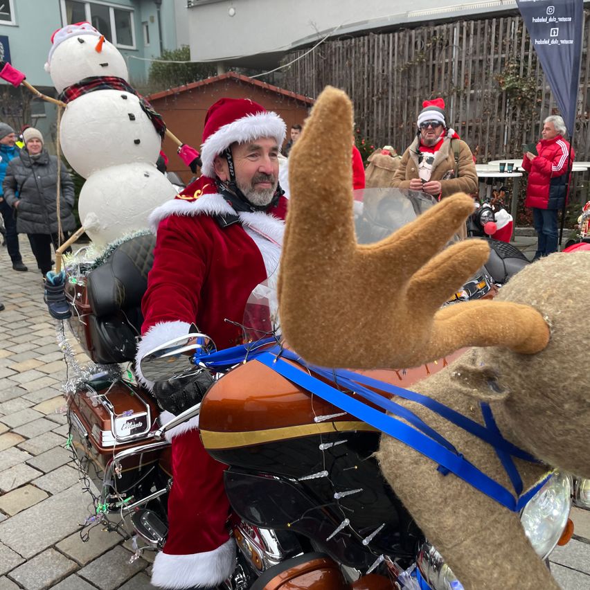 Ein Mann in Santa-Claus-Kleidung fährt ein Motorrad mit Schneemann- und Rentierdekoration, umgeben von Menschen in festlicher Kleidung.