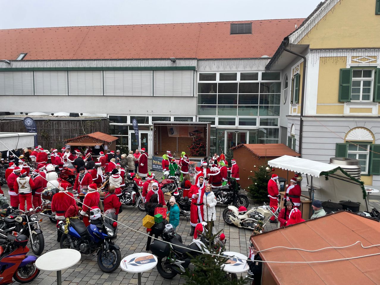 Eine große Menschenmenge, die als Weihnachtsmann verkleidet ist, versammelt sich vor einem Gebäude mit rotem Dach und Glasfenstern um Motorräder herum, während einer festlichen Veranstaltung.