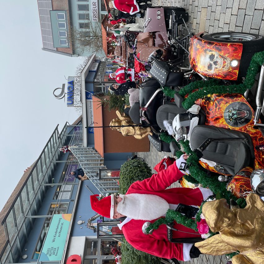 Der Weihnachtsmann steht vor einer Reihe von Motorrädern, die geparkt und für die Feiertage geschmückt sind. Sie befinden sich vor einem Gebäude mit dem Schild 'UNIGA'.
