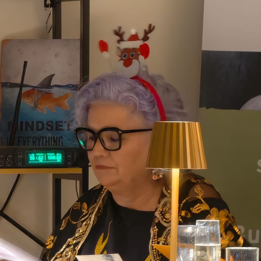 Eine Frau sitzt vor einem Schreibtisch mit einer Lampe, trägt eine Brille und eine Haarspange mit Rentierohren. Hinter ihr befindet sich ein Poster, das einen Hai und das Wort 'Mindset' zeigt. Rechts ist ein verschwommenes Bild eines grünen Banners mit weißem Text zu sehen.