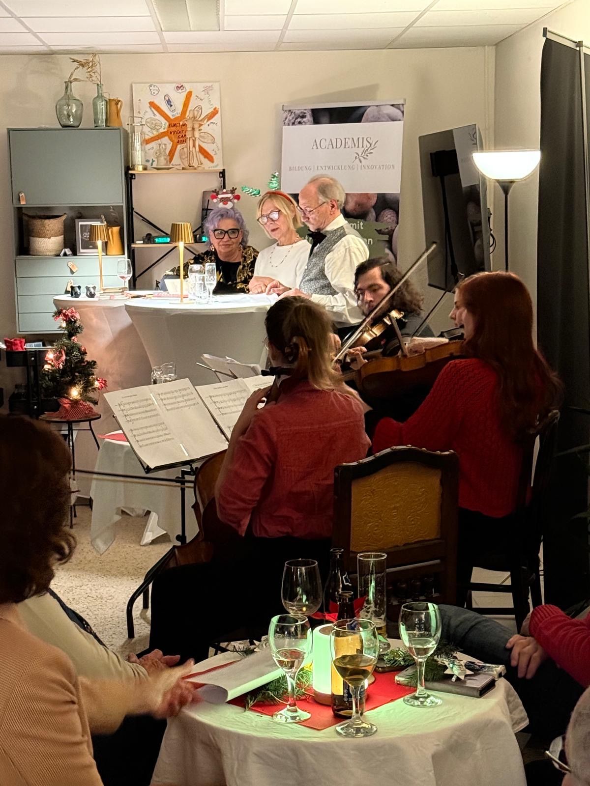 Eine Gruppe von Menschen befindet sich in einem Raum und hört eine Aufführung. Ein Geiger und ein Cellist spielen sitzend. Vor ihnen steht ein Tisch mit Notenständern und Weingläsern.