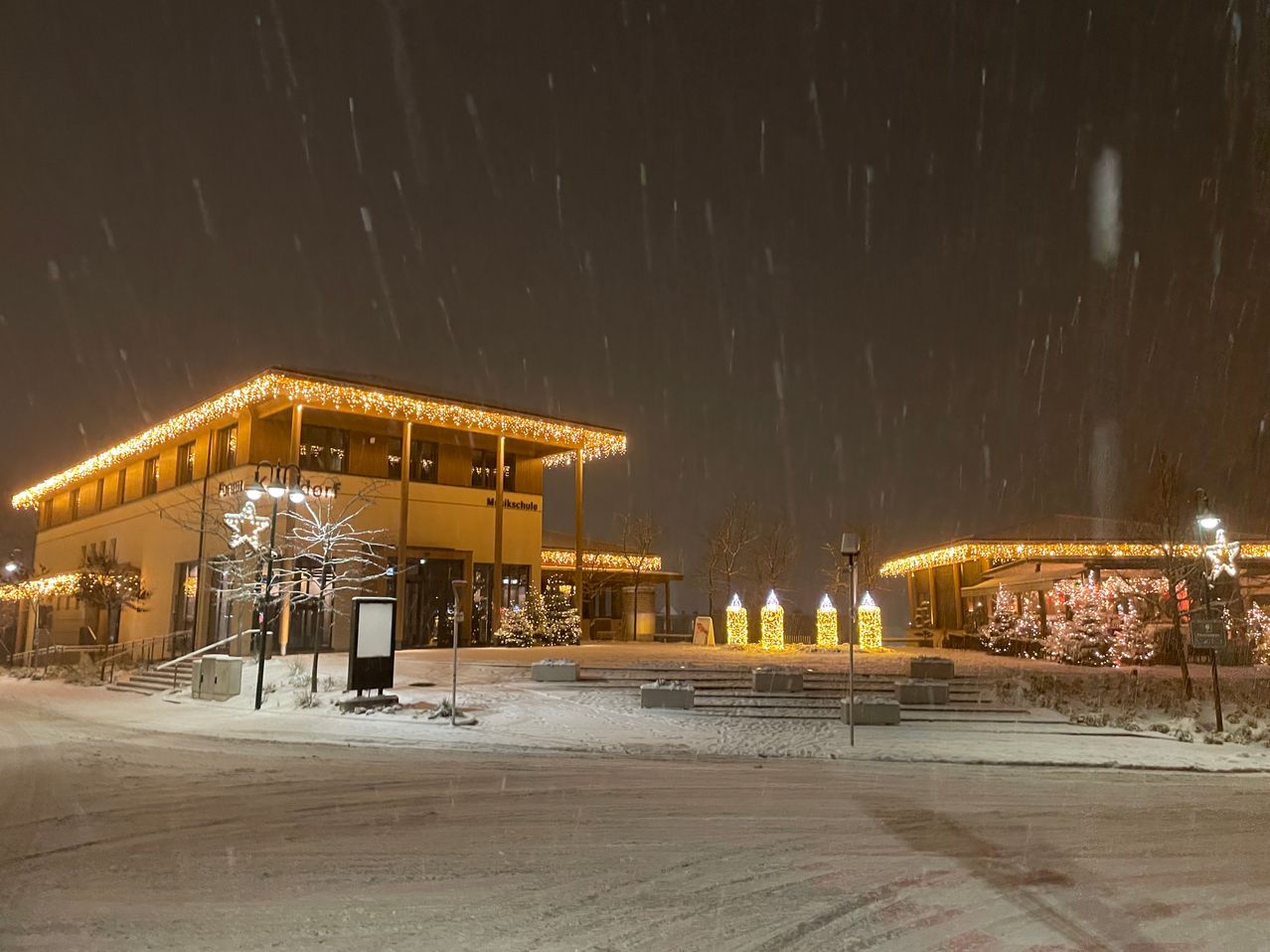 Ein Gebäude mit Weihnachtsdekoration und Lichtern ist in der Nacht im Schnee zu sehen. Der Bereich ist von Schnee umgeben und im Vordergrund stehen Weihnachtsbäume.
