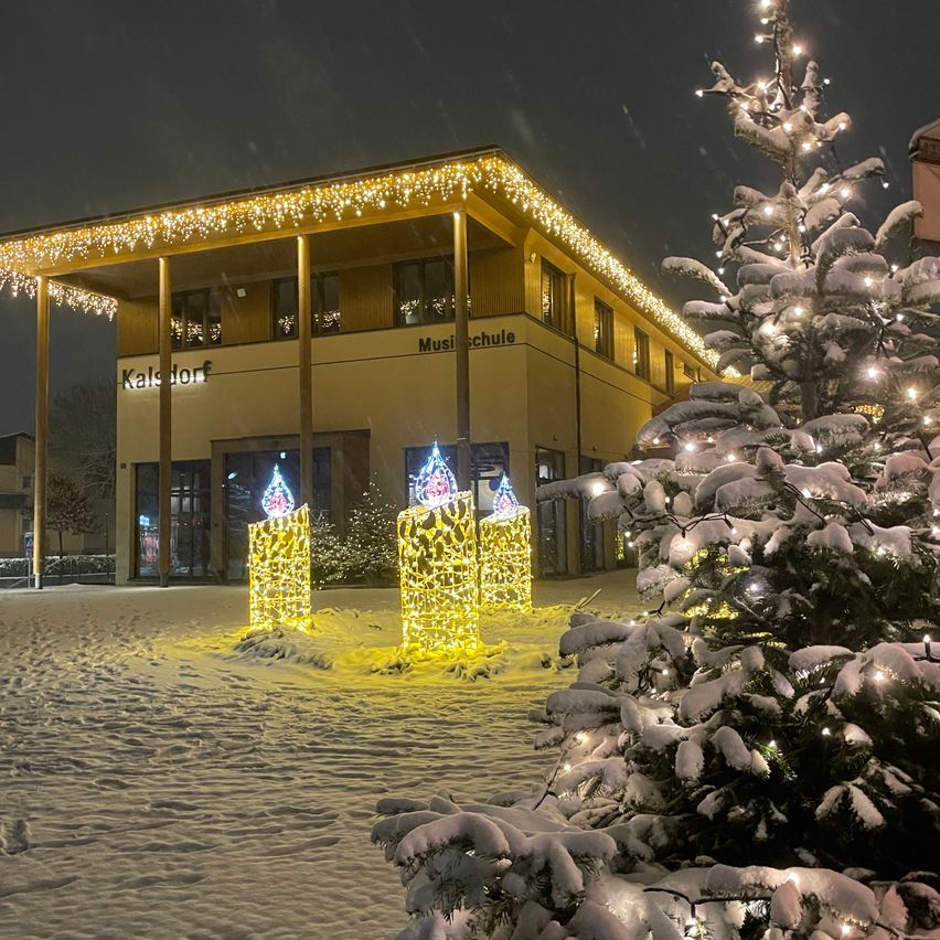 Ein schneebedecktes Gebäude mit Weihnachtsbeleuchtung und einem festlichen Baum steht in einer verschneiten Landschaft. Das Gebäude heißt Kalsdorf.