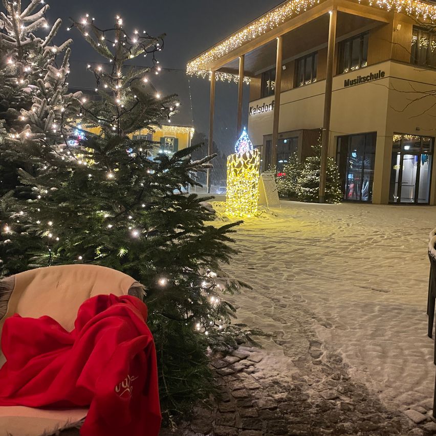 Eine verschneite Szene mit einem beleuchteten Weihnachtsbaum, einer leuchtenden Weihnachtsdekoration und einem Mantel, der über einen Stuhl vor einem Gebäude mit den Worten Kalsdorf und Musikschule geworfen wurde.