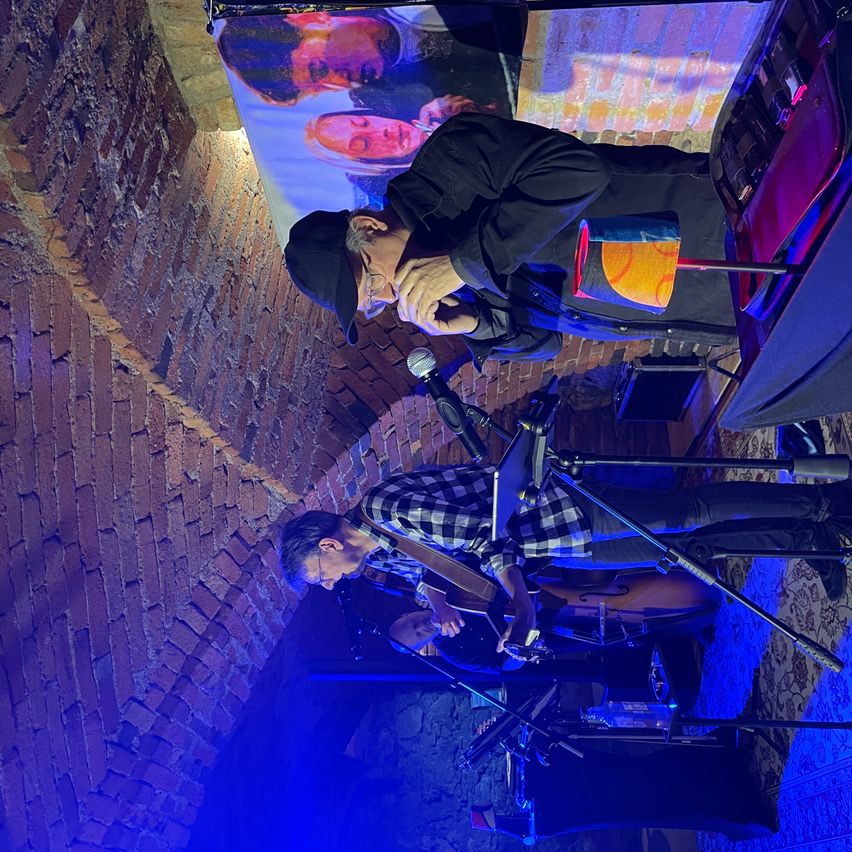 Eine Band spielt unter einem blauen Licht. Der Leadsänger spielt die Mundharmonika. Ein Mann spielt Gitarre und ein anderer Cello.