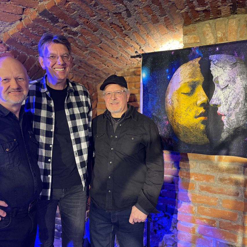 Drei Männer stehen vor einer Ziegelwand. Der Mann in der Mitte trägt eine Brille und einen Hut. Dahinter hängt ein Bild mit zwei Gesichtern, eines gelb und eines weiß.