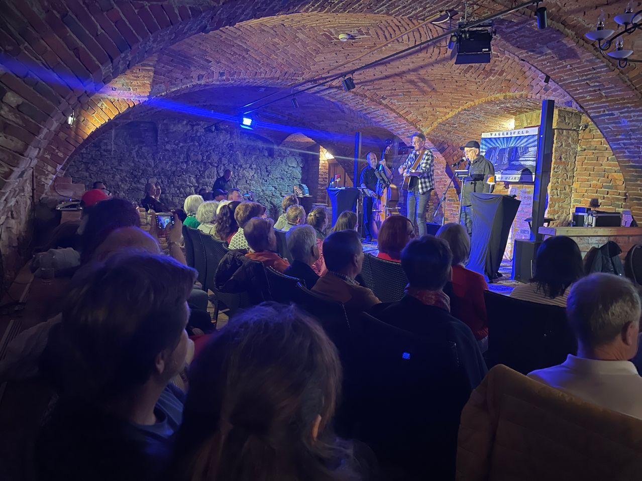 Eine Gruppe Musiker tritt auf der Bühne in einem schwach beleuchteten Raum aus Ziegelsteinen auf. Das Publikum, auf Stühlen sitzend, hört aufmerksam zu. Eine blaue Scheinwerfer beleuchtet die Bühne.