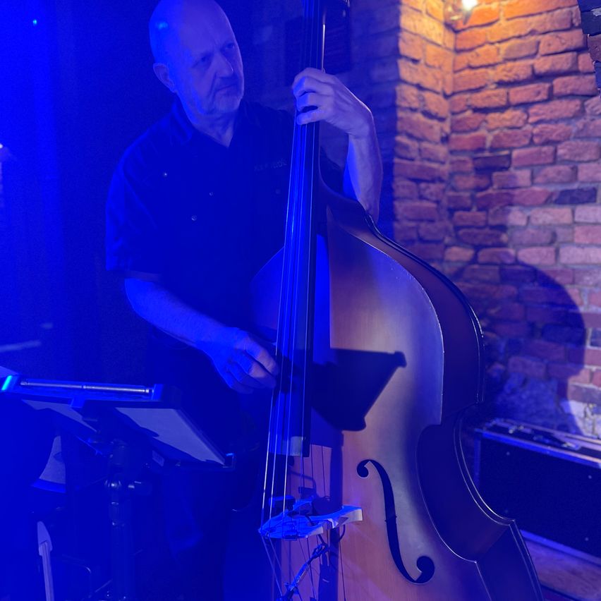 Ein älterer Mann spielt ein Bassgitarre, beleuchtet von blauen Lichtern, in einem schwach beleuchteten Raum mit einer Ziegelwand.