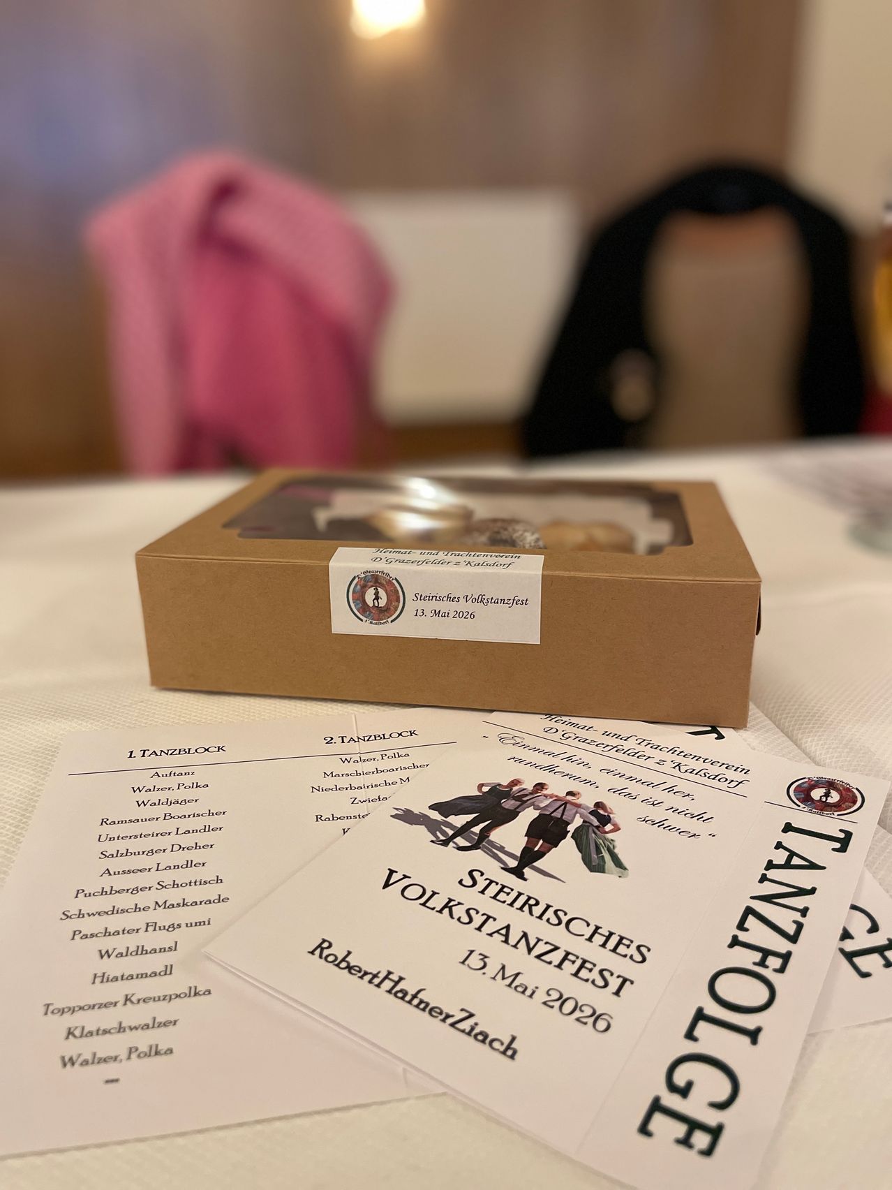 A box of traditional sweets sits on a table next to a pamphlet advertising the Steirisches Volkstanzfest, a dance festival in Austria, with a blurry pink cloth in the background.