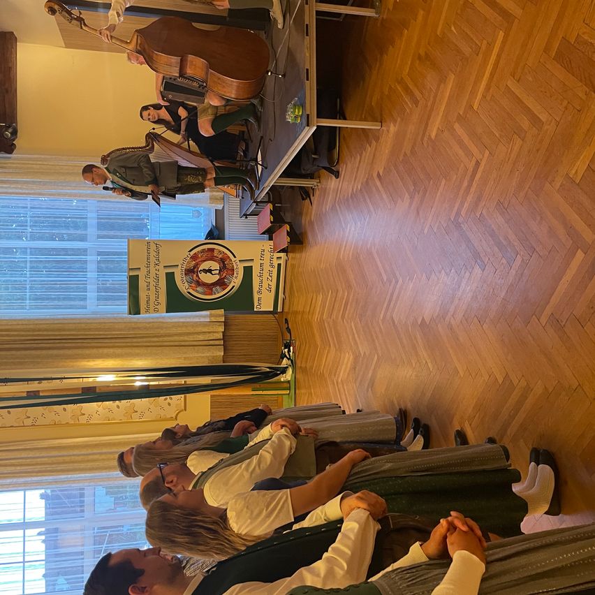 An indoor musical performance with people in traditional clothing. A man plays a harp while another holds a cello. Several people are seated and listening. A sign with green and white text is in the background.