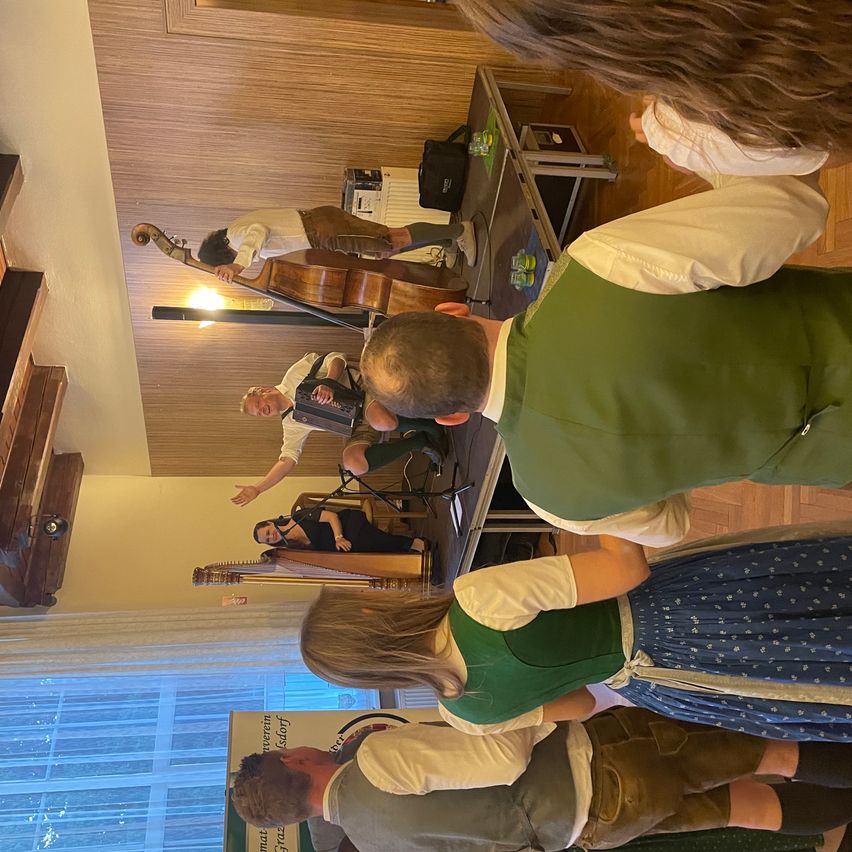 A group of people dressed in traditional clothing are watching a musician play the cello and accordion, with a harpist in the background. The setting appears to be a cultural event.