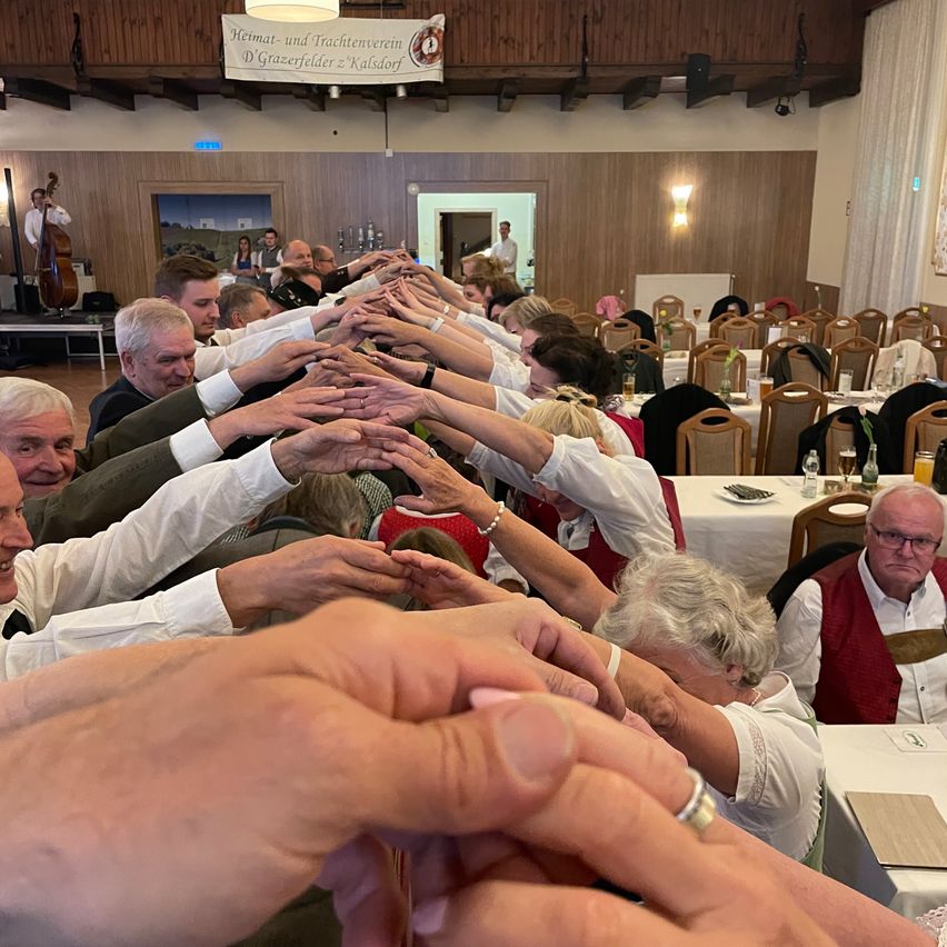 A group of people in a hall hold hands together in a circle, possibly at a gathering. A banner hangs above them.