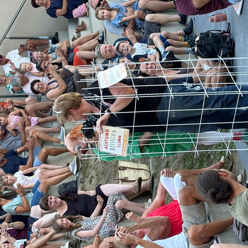 Eine Menschenmenge sitzt auf den Stufen, einige lächeln und machen Fotos. Eine Person in einem schwarzen Shirt steht in einem Metallkäfig mit einem Schild, das 'Achtung Bissiger Clown' besagt.