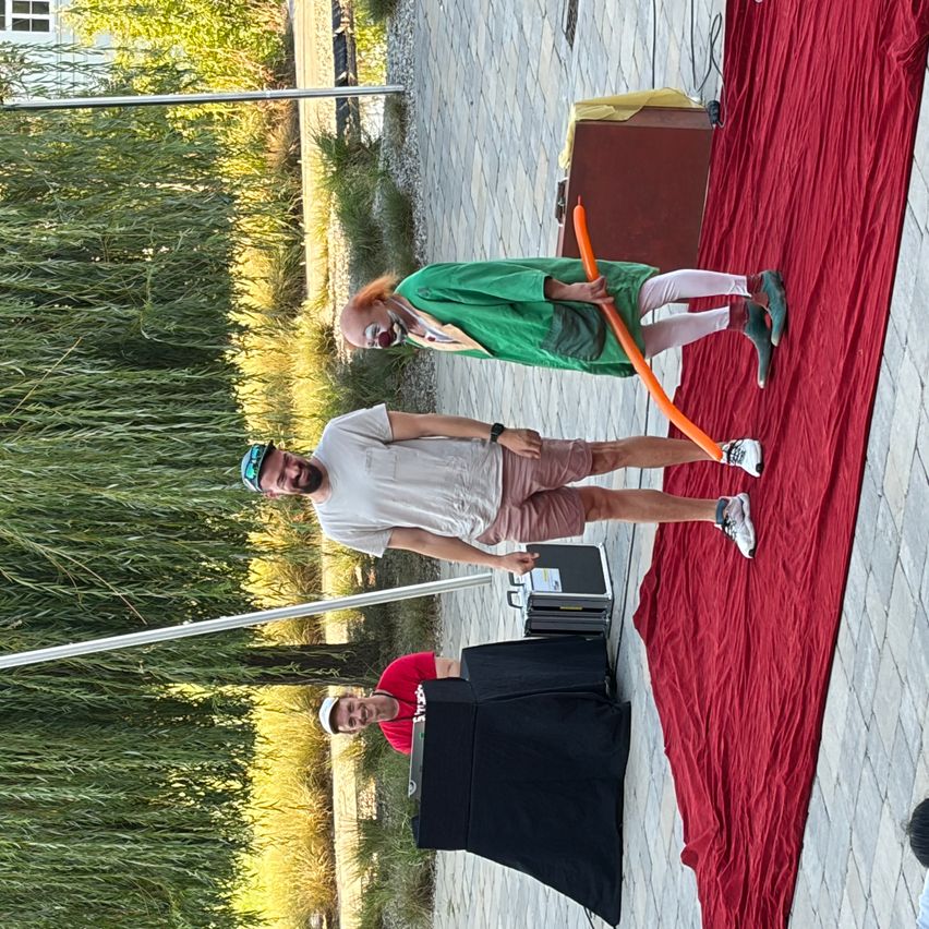 Zwei Männer und ein Clown stehen auf einem roten Teppich mit rotem Hintergrund. Ein Mann hält einen orangefarbenen Luftballon.