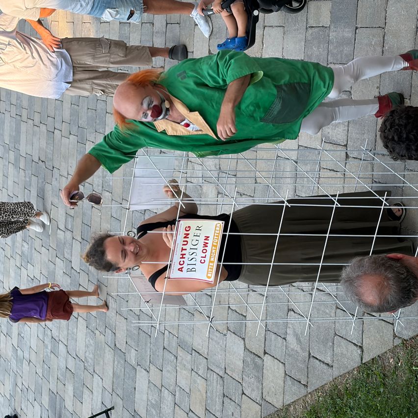 Eine Person, die als Clown verkleidet ist, liegt auf einem Drahtgitterzaun, während eine andere Person ein Schild mit dem Text 'ACHTUNG BISSIGE CLOWNER' hält. Eine Frau steht hinter dem Zaun und ein Kind steht links. Ein Kinderwagen steht hinter ihnen.
