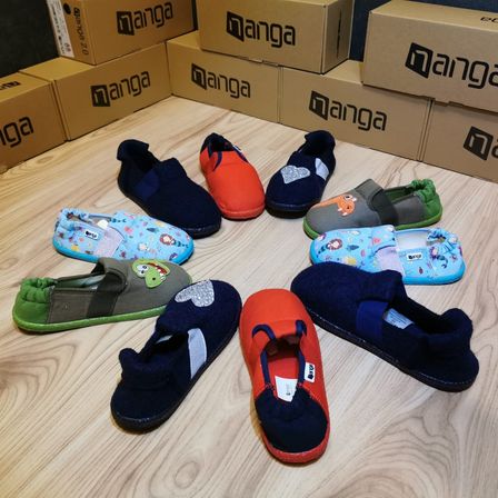 Mehrere Kinderhausschuhe in verschiedenen Farben sind in einem Kreis auf einem Holzboden angeordnet, mit Manga-Kisten im Hintergrund.