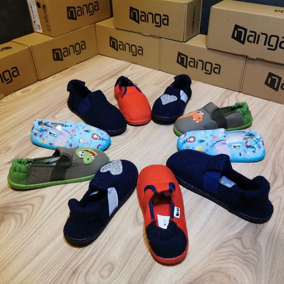 Mehrere Kinderhausschuhe in verschiedenen Farben sind in einem Kreis auf einem Holzboden angeordnet, mit Manga-Kisten im Hintergrund.