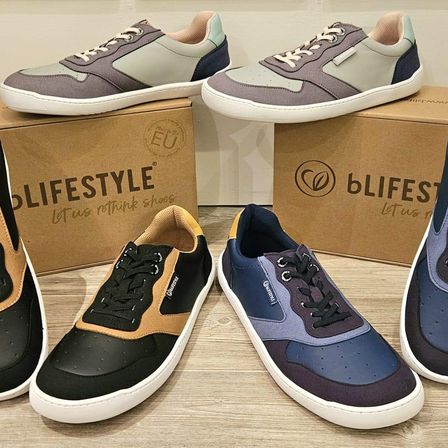 Fünf Paar Sneaker auf einer Holzoberfläche, drei mit weißen Sohlen und zwei mit farbigen Sohlen. Zwei Kartons mit den Aufschriften 'Lifestyle' und 'bLifeStyle' im Hintergrund.