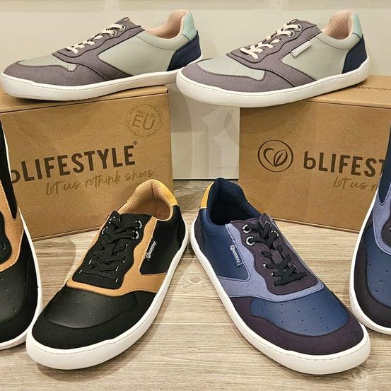 Fünf Paar Sneaker auf einer Holzoberfläche, drei mit weißen Sohlen und zwei mit farbigen Sohlen. Zwei Kartons mit den Aufschriften 'Lifestyle' und 'bLifeStyle' im Hintergrund.