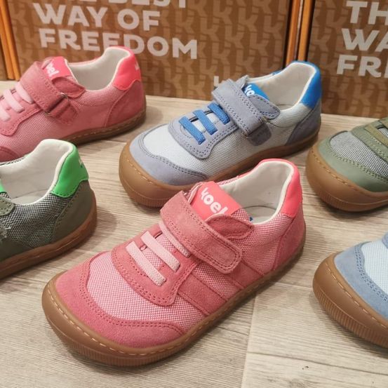 Mehrere Paare Kinderschuhe in verschiedenen Farben sind auf einer Holzoberfläche ausgestellt. Die Schuhe haben Riemen und Schnürsenkel zum einfachen Anziehen. Im Hintergrund sind Kisten mit dem Text 'Way of Freedom' zu sehen.