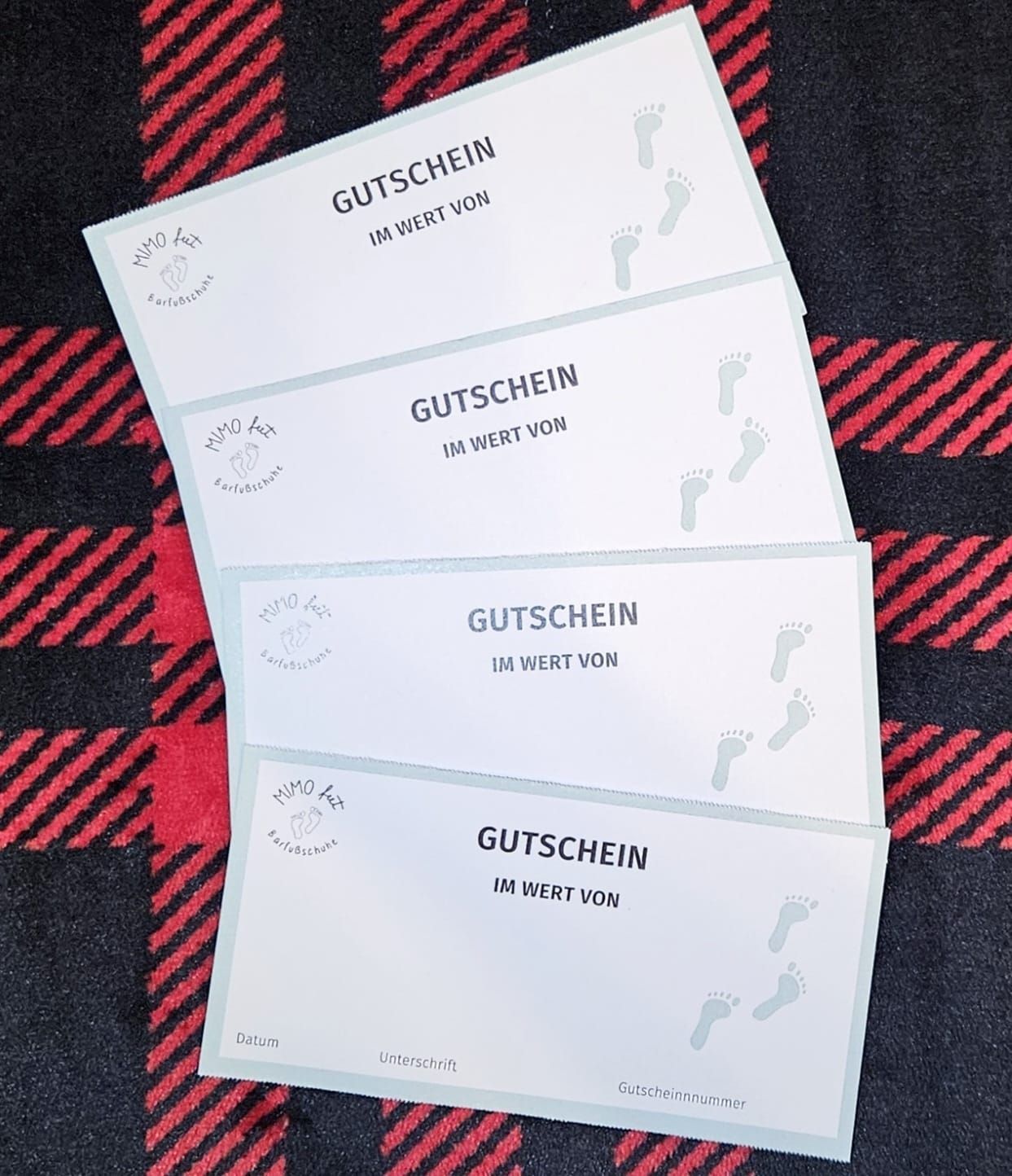 Vier weiße Geschenkgutscheine mit einem Baby-Fußabdruck auf einem rot-schwarzen Tuch. Der Text lautet 'Gutschein' und 'Im Wert von'.