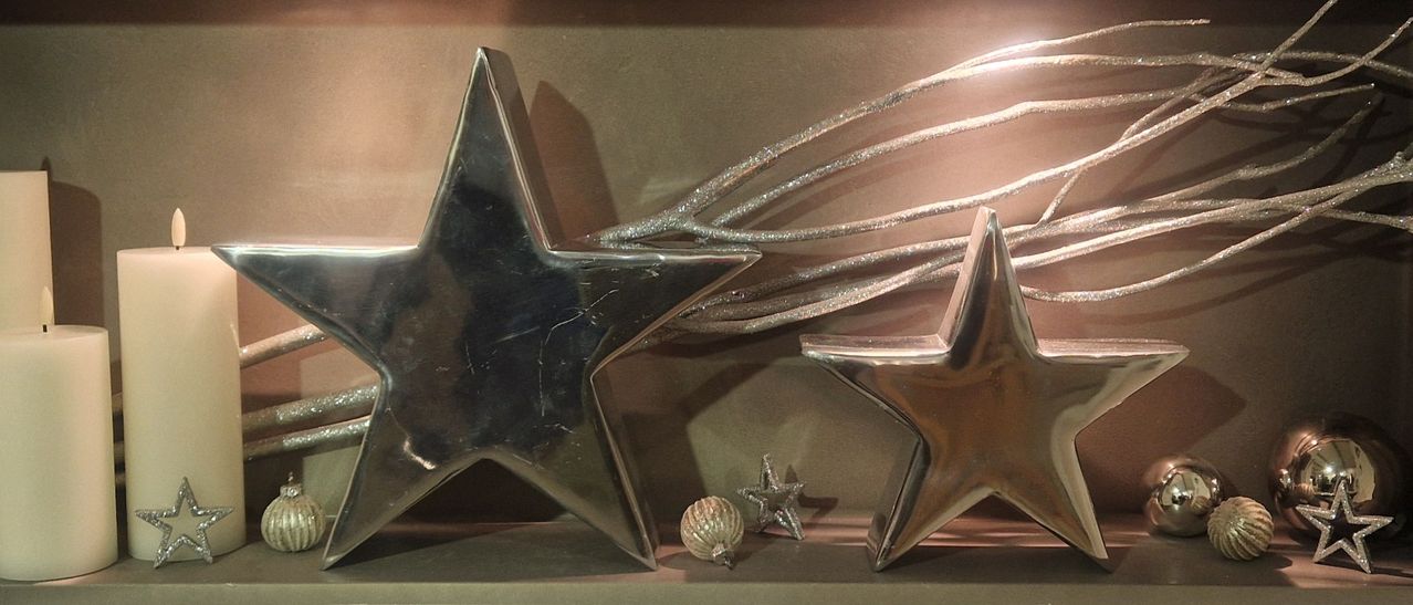 Eine glänzende metallische Sternendekoration wird mit zwei kleineren Sternen und einer Muschel ausgestellt. Der metallische Stern ist silber und hat eine reflektierende Oberfläche. Dahinter befinden sich einige silberne Bänder.