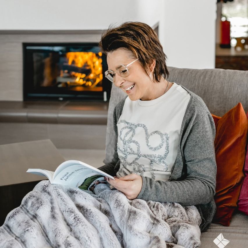 A woman sits on a couch, smiling, holding a book. She wears glasses and a gray cardigan. Behind her, a fireplace is lit.