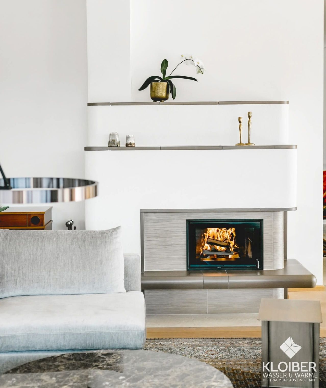 A modern living room with a fireplace, white walls, and shelves. A gray couch with a pillow sits nearby, and a plant and two candles are on the shelves.