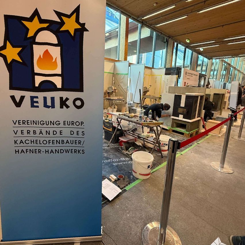 A busy area with a banner for VEUKO, a European association of tile installers, has people working on various tasks with tools and materials scattered around.