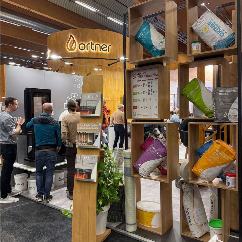 People are gathered at an indoor exhibit, observing a display of bags, buckets, and a plant. Behind them, an 'ortner' logo is displayed on a wooden wall. Some individuals are seated at a table.