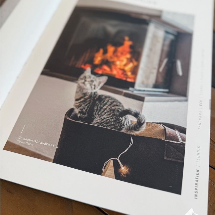 Ein Fotobuch zeigt eine Katze, die auf einem Kissen neben einem Kamin sitzt. Das Bild zeigt helle Flammen und ist auf hochwertigem Papier gedruckt.