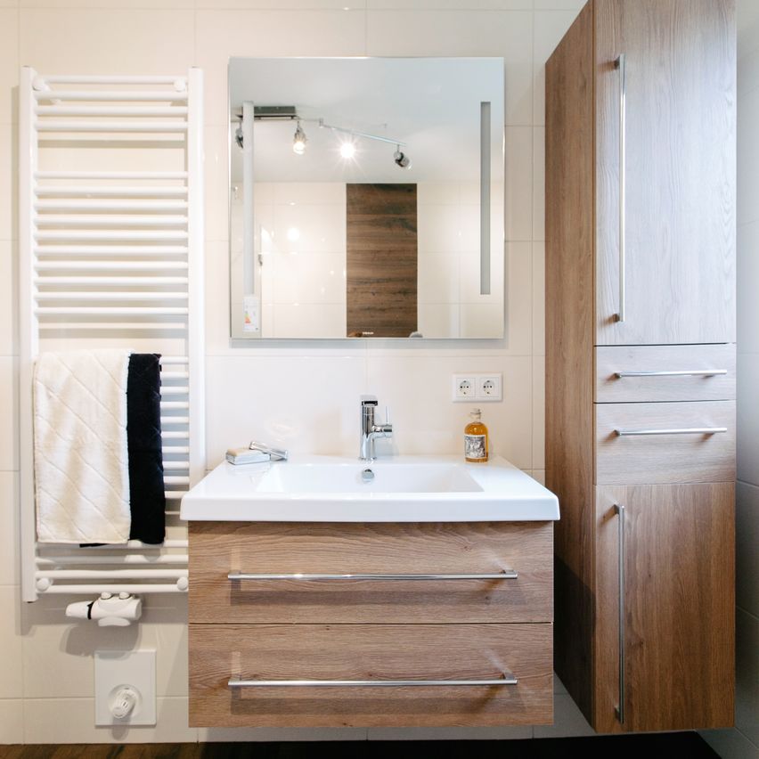 Ein modernes Badezimmer mit einem weißen Waschbecken, Holzschränken und einem Spiegel, der eine geflieste Wand und einen Handtuchhalter reflektiert.